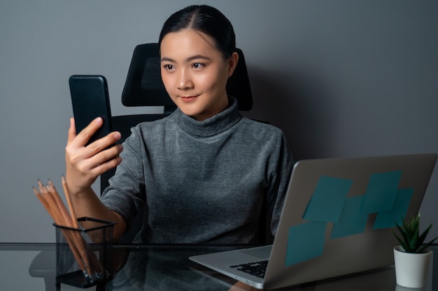 Glückliches Lächeln der asiatischen Frau unter Verwendung des Smartphones und Arbeiten an einem Laptop im Büro