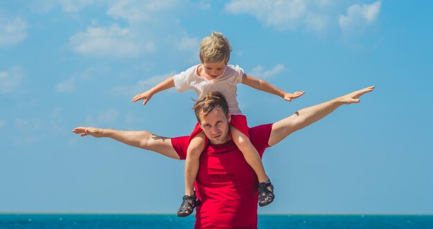 Glückliches Lächeln Vater Umarmungen hält auf den Schultern Sohn Junge männliches Kind stehen blau lazur Meer Panorama Skyline Horizont im Sonnenschein Tag klarer Himmel Spielshow ausgestreckte Hände wie Flugzeug Reisebeziehung