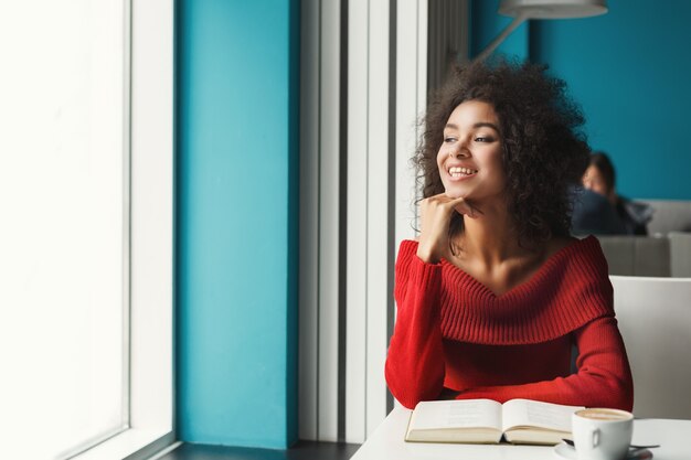 Glückliches lächelndes afroamerikanisches Mädchen am Cafétisch. Junge Frau, die heißen Kaffee genießt und Lieblingsbuch liest, Fenster betrachtend. Freizeit, Bildung und urbanes Lifestyle-Konzept.