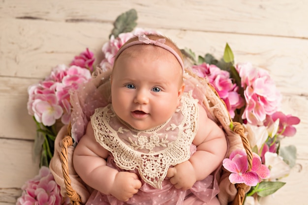 Glückliches lächelndes Baby in einem rosa Kleid liegt auf ihrem Rücken in einem Blumenkorb