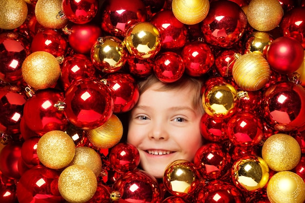 Glückliches lächelndes Kind mit Weihnachtskugel-Spielwaren. Kind über rotem und goldenem Flitter-Hintergrund. Glänzende helle Weihnachtsbaum-Feiertagsdekoration