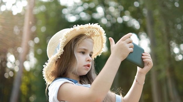 Glückliches lächelndes Kindermädchen, das im Sommer draußen im Handy schaut