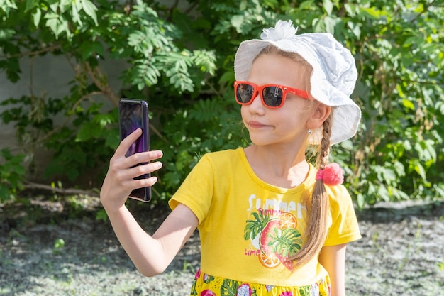 Glückliches lächelndes Kindermädchen, das im Sommer im Freien im Handy schaut Ein fröhliches kleines Mädchen in einem gelben Kleid