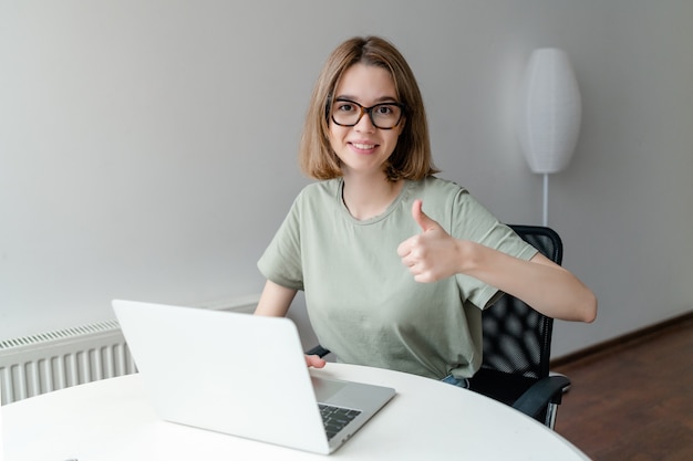 Glückliches lächelndes Mädchen, das online arbeitet oder studiert und lernt, während Notebook verwendet wird, das Daumen hoch zeigt. Freiberufliches Arbeitskonzept