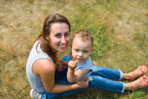 Glückliches lächelndes Mutter- und Sohnkind, das auf Gras am Sommertag sitzt