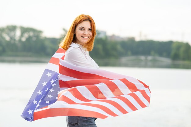 Glückliches lächelndes rothaariges Mädchen mit USA-Nationalflagge auf ihren Schultern