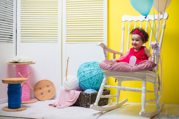 Glückliches lächelndes süßes Baby, das auf Lehnsessel mit Geburtstagsballonen sitzt