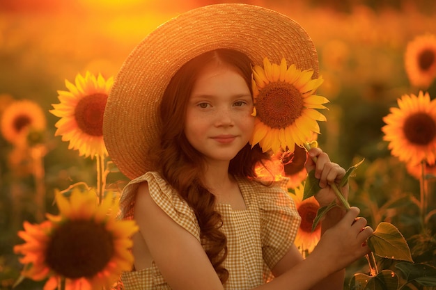 Glückliches Leben im Sommer Porträt eines schönen rothaarigen Mädchens mit Hut auf einem Sonnenblumenfeld mit Blumen in den Händen in den Strahlen der untergehenden Sonne