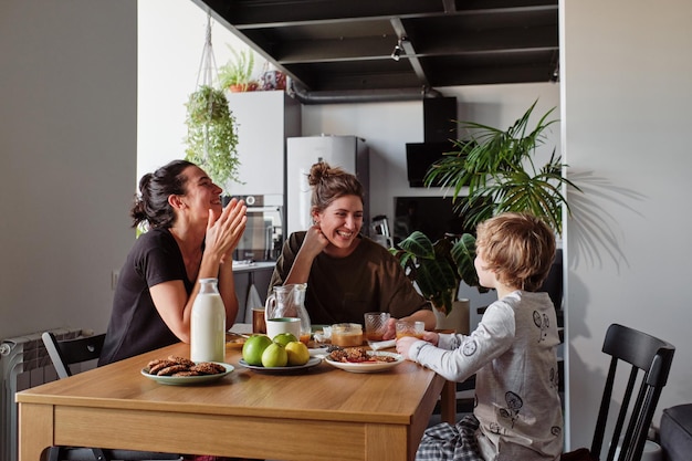 Glückliches lesbisches Paar lacht beim Frühstück am Tisch, während ihr kleiner Sohn lustige Witze redet