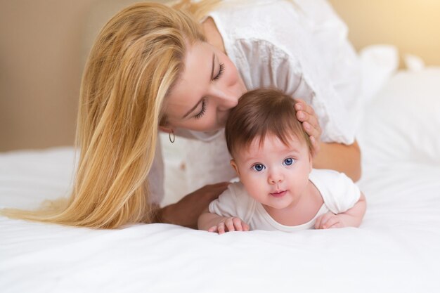 Glückliches liebevolles Familienkonzept. Schöne Mutter, die mit ihrem Baby im Schlafzimmer spielt. Sie lächeln und umarmen sich zusammen auf weißer Bettwäsche