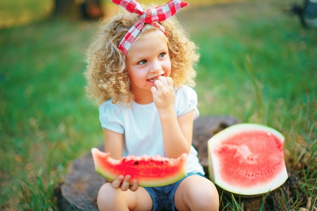 Glückliches lockiges Mädchen, das auf Stumpf sitzt und Wassermelone draußen im Frühlingspark isst