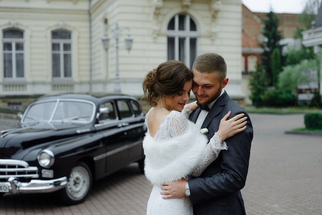 Glückliches Luxushochzeitspaar, das nahe Retro mit Blumenstraußauto im Herbst küsst und umarmt