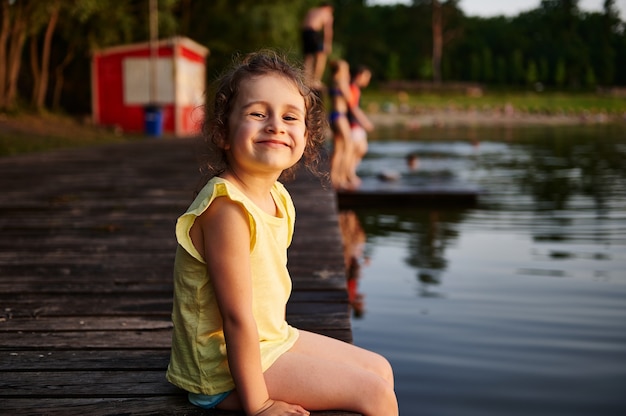 Glückliches Mädchen auf einem Pier lächelt, wenn man die Kamera betrachtet. Freizeitkonzepte im Sommer. Hitzesommer, Sommerabende in der Natur, Zeit an der frischen Luft verbringen