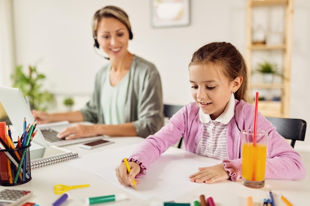 Glückliches Mädchen, das auf Papier zeichnet, während ihre Mutter zu Hause arbeitet