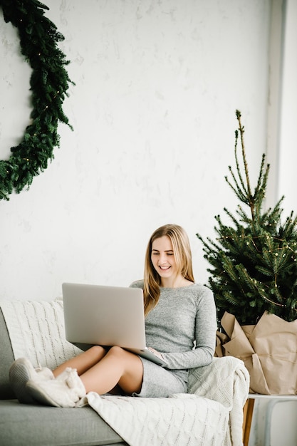 Glückliches Mädchen, das Freunde oder Familie mit Neujahr im Video-Chat auf dem Laptop begrüßt Junge Frau mit Laptop zu Hause im Büro in der Nähe von geschmückter Kiefer zu Hause mit weihnachtlichem Interieur mit Kopierraum