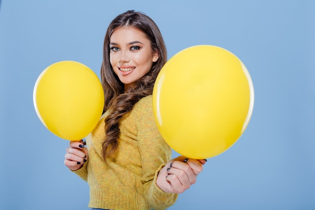 Glückliches Mädchen hält gelbe Luftballons und lächelt für die Kamera, während es Spaß gegen isoliert auf bl ...