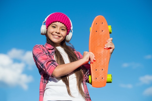 Glückliches Mädchen hält Penny Board, das Musik im sonnigen Himmel im Freien Transport mit Kopfhörern hört