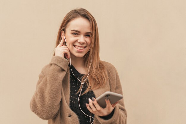 Glückliches Mädchen hört Musik, hält ein Smartphone in ihren Händen, trägt einen braunen Mantel, der auf beigem Hintergrund isoliert wird