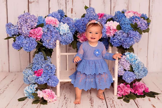 Glückliches Mädchen im blauen Kleid sitzt auf Bett mit blauen und rosa Hortensienblumen