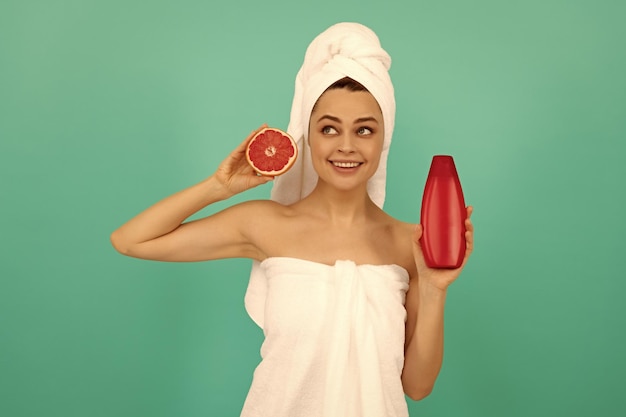 Glückliches Mädchen im Handtuch mit Grapefruit-Shampoo-Flasche auf blauem Hintergrund