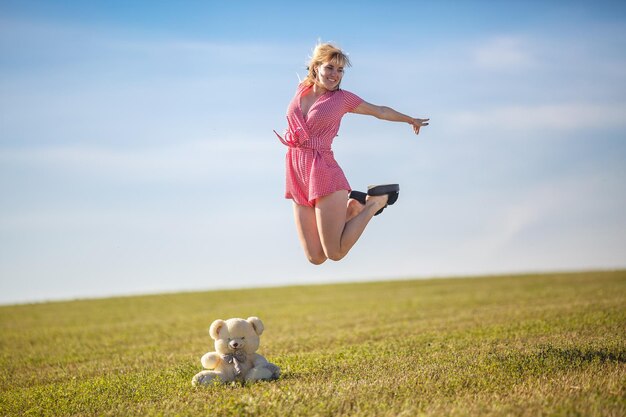 Glückliches Mädchen im karierten Anzug, das auf dem Feld in der Nähe des Teddybären springt