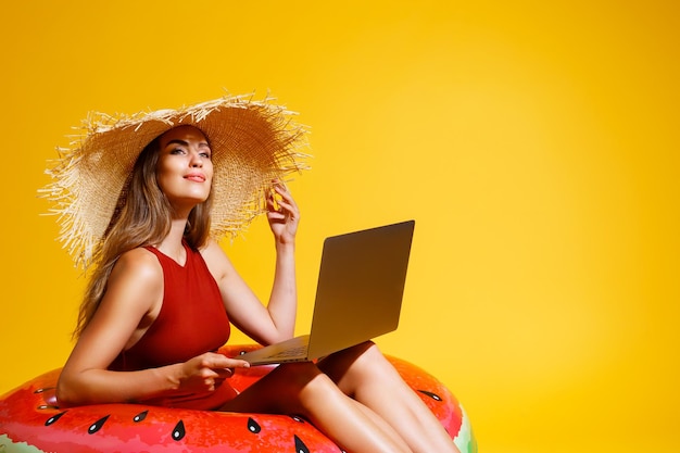 Glückliches Mädchen im Stroh sitzt in aufblasbarem Ring arbeitet am Laptop auf gelbem Hintergrund Freiberuflerreisen