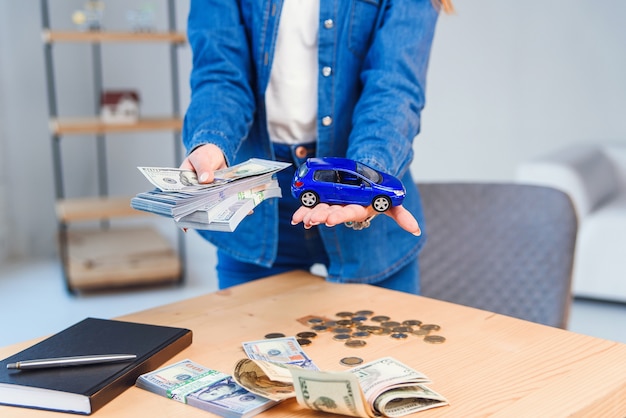 Glückliches Mädchen in Brille hält eine Geldsumme in einer Hand und kleines Modell ihres zukünftigen Autos in einer anderen Hand.