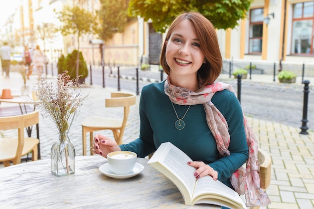 Glückliches Mädchen in einem Café im Freien liest ein Buch und trinkt Kaffee