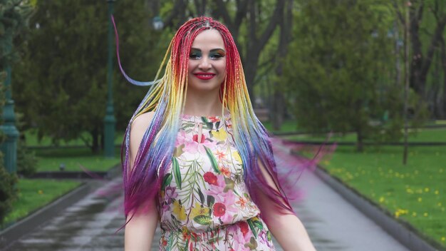 Glückliches Mädchen in einem Kleid mit afrikanischen Zöpfen des Regenbogens und hellem buntem Make-up. Süß lächelt und wirft ihre Haare hoch und wirbelt bei einem Spaziergang im Frühlingspark herum