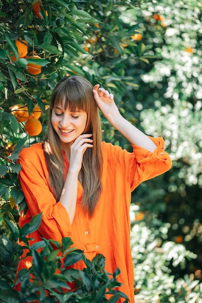 Glückliches Mädchen in orangefarbenem Kleid posiert vor der Kamera, indem es nach unten schaut und ihre Hände im orangefarbenen Garten hochhält