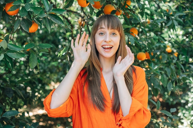 Glückliches Mädchen in orangefarbenem Kleid schaut in die Kamera, indem sie ihre Hände im orangefarbenen Garten erhebt