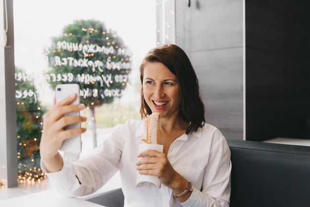 Glückliches Mädchen macht ein Selfie auf einem Handy, während es in einem Café einen Twister isst.