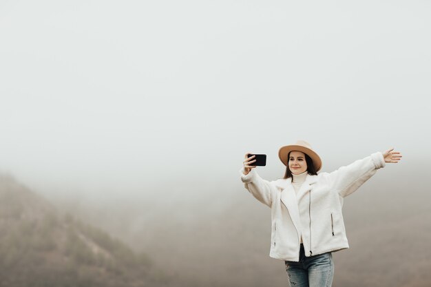 Glückliches Mädchen mit der Hand, die selfie auf Klippenrand des Berggipfels in der nebligen Zeit nimmt.