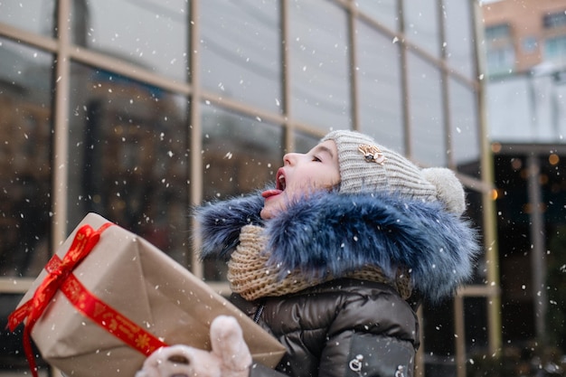 Glückliches Mädchen mit einer Geschenkbox zu Weihnachten fängt Schneeflocken mit seiner Zunge in einer Stadt im Winter mit Schnee auf einem festlichen Markt mit Dekorationen und Lichtern. Warme Kleidung, Strickmütze, Schal und Pelz.