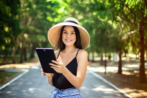 Glückliches Mädchen mit einer Tablette in der Hand und einem Hut, einem Geschäft und einer sozialen Tätigkeit im Internet tragend, die in der Natur sind.