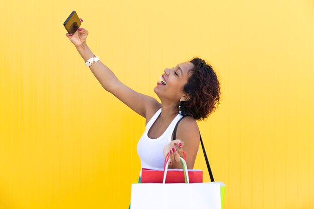 Glückliches Mädchen mit Einkaufstüten nimmt Selfie auf ihrem Telefon an einer gelben Wand.