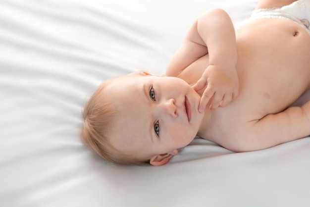 Glückliches nacktes Baby in Windeln, das auf dem Rücken auf dem weißen Laken liegt und sein Gesicht berührt. Babymädchen mit roten Haaren, die in die Kamera blicken. Heller Hintergrund