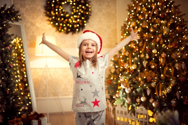 Glückliches nettes mädchen in den weihnachtspyjamas, sankt-helferhut hob ihre hände oben an.