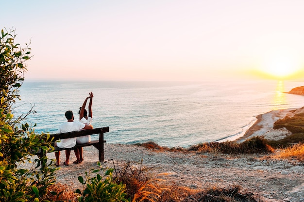 Glückliches Paar bei Sonnenuntergang. Mann und Frau im Morgengrauen. Paar sitzt auf einer Bank am Meer. Verliebtes Paar, das sich am Meer küsst. Mann und Frau reisen. Flitterwochen. Sonnenuntergang am Meer. Morgendämmerung am Meer