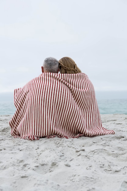 Glückliches paar, das auf dem sand mit decke um sie sitzt