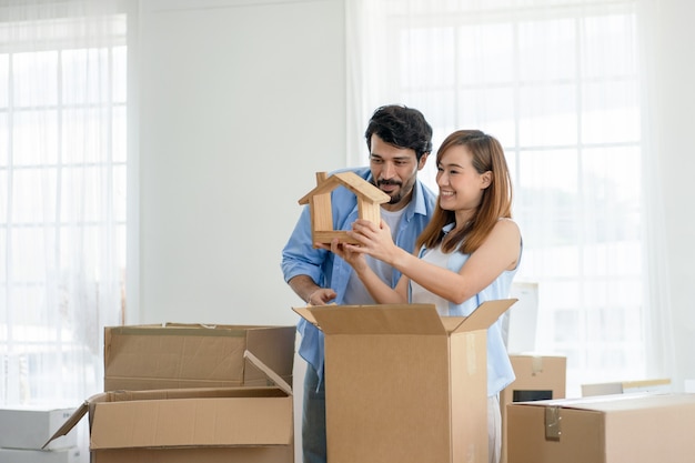 Glückliches Paar, das hölzernes Hausmodell hält, während Kisten für Umzug in ein neues Haus packen