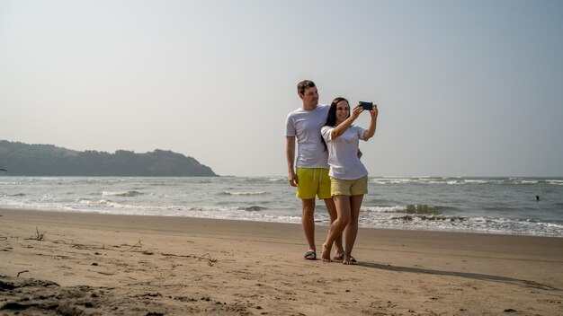 Glückliches Paar, das Selfies in der Nähe des Meeres macht Liebespaar, das sich während des Dates am Strand gegen das wehende Meer und den wolkenlosen Himmel umarmt