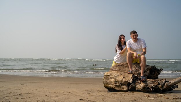 Glückliches Paar, das sich auf Treibholz in der Nähe des Meeres ausgibt Liebendes Paar, das sich während des Dates am Strand gegen das wehende Meer und den wolkenlosen Himmel umarmt