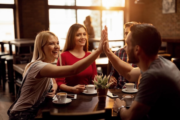 Glückliches Paar, das sich gegenseitig Highfive gibt und Spaß hat, während es sich mit Freunden in einem Café trifft. Der Fokus liegt auf der Frau