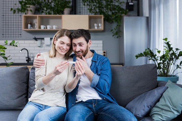 Glückliches Paar, das zu Hause auf dem Sofa sitzt, Mann und Frau bekamen einen positiven Schwangerschaftstest
