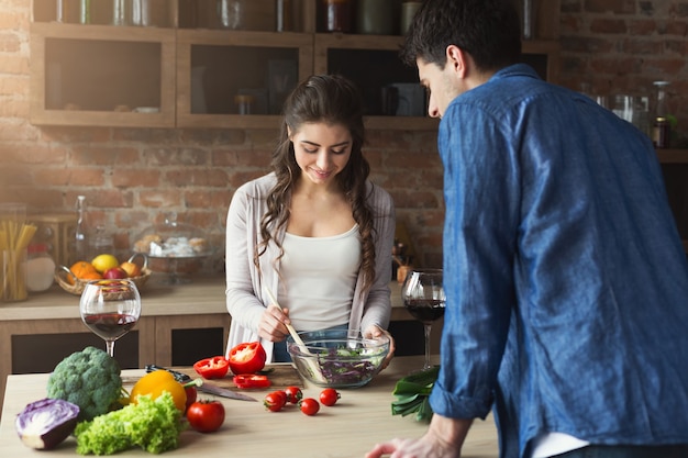 Glückliches Paar, das zu Hause gesundes Essen in ihrer Loft-Küche kocht und probiert. Frau und Mann trinken Wein. Gemüsesalat zubereiten.