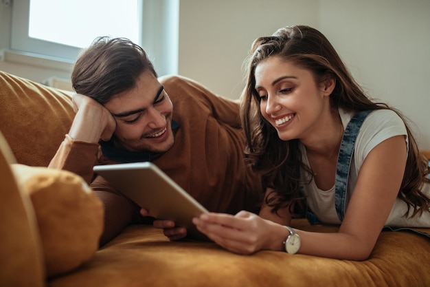 Glückliches Paar, das zu Hause mit einem digitalen Tablet auf dem Sofa liegt