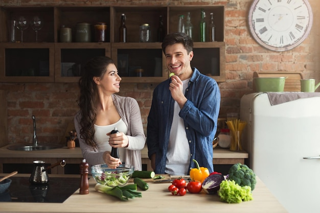 Glückliches Paar, das zusammen gesundes Essen kocht, Mann isst Gurke in ihrer Dachbodenküche zu Hause. Gemüsesalat zubereiten.