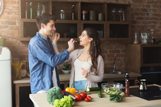 Glückliches Paar, das zusammen gesundes Essen kocht und Gurken in ihrer Dachbodenküche zu Hause isst. Gemüsesalat zubereiten.