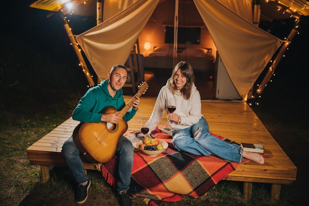 Glückliches Paar entspannt sich im Glamping am Herbstabend, trinkt Wein und spielt Gitarre in der Nähe eines gemütlichen Lagerfeuers Luxus-Campingzelt für Erholung und Erholung im Freien Lifestyle-Konzept